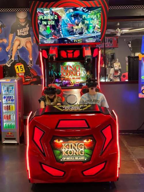 Two players wearing VR headsets at a King Kong arcade game, with a vibrant red cabinet and a screen above displaying gameplay.