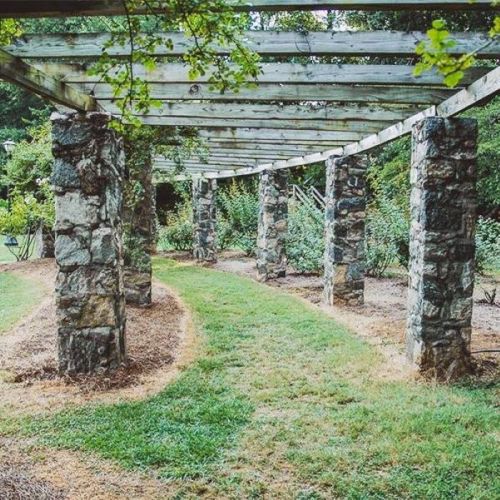 A stone pergola with wooden beams, surrounded by lush greenery and a winding path in a serene garden setting.