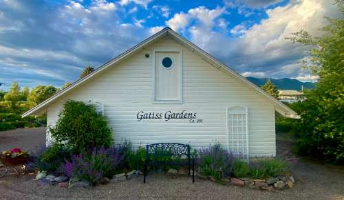 A white house with "Gaitss Gardens" sign, surrounded by greenery and flowers under a blue sky with clouds.