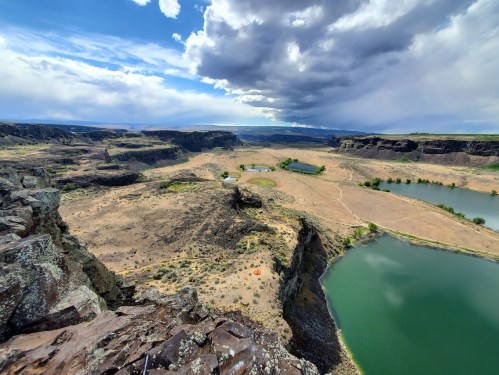 A panoramic view of a rugged landscape with rocky cliffs, green lakes, and a dramatic sky filled with clouds.
