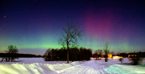 A snowy landscape under a vibrant aurora borealis, with trees and a distant building illuminated by the colorful sky.