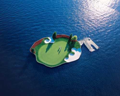 Aerial view of a small golf green island surrounded by water, featuring sand traps and a dock.