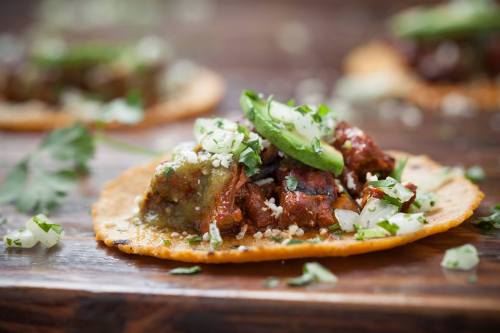 Close-up of a taco topped with meat, green sauce, sliced cucumbers, onions, and cilantro on a wooden surface.