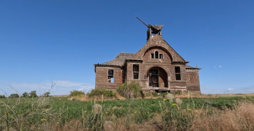 Abandoned wooden building with a peaked roof, surrounded by tall grass and under a clear blue sky.