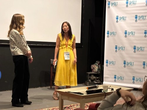 Two women on stage at a film festival, one in a yellow dress speaking, the other listening. A banner in the background.