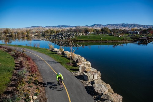 A cyclist rides along a path by a calm lake, surrounded by trees and mountains under a clear blue sky.