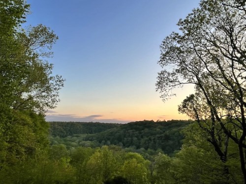 A serene landscape at sunset, featuring lush green trees and rolling hills under a clear sky.