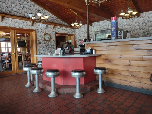 A cozy diner interior featuring a circular counter with stools and rustic stone walls. Warm lighting enhances the atmosphere.