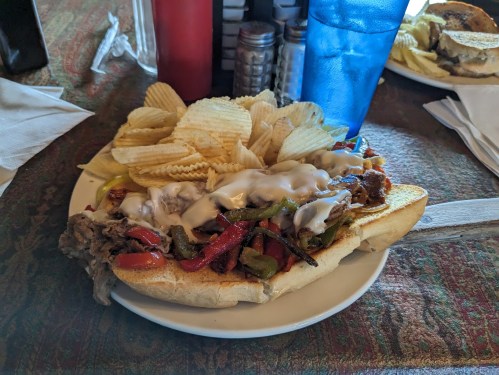 A loaded sandwich with grilled peppers and onions, topped with mayonnaise, served with a side of potato chips.