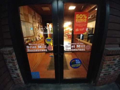Double glass doors of Crist Mill Restaurant with promotional signs and a New York Lottery sticker.