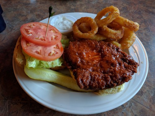 A plate featuring a spicy chicken sandwich with lettuce, tomato, pickles, onion rings, and a side of dipping sauce.