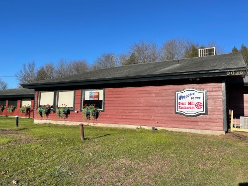 A red wooden building with a sign reading "Welcome to the Grist Mill Restaurant" and flower boxes under the windows.