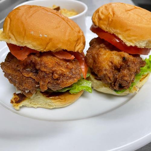 Two crispy chicken sandwiches with lettuce and tomato on soft buns, served with a side of dipping sauce.