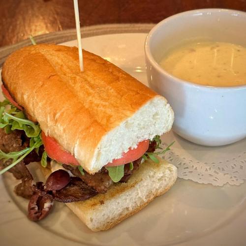 A sandwich with meat, lettuce, and tomato on a plate, accompanied by a small bowl of creamy soup.