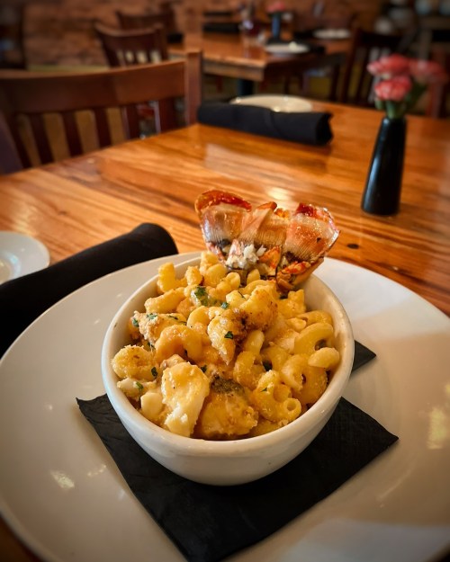 A bowl of creamy macaroni and cheese topped with lobster, served on a black napkin at a restaurant table.