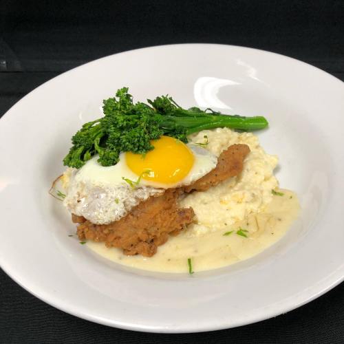 A plate of fried chicken topped with a sunny-side-up egg, served with creamy grits and sautéed broccolini.