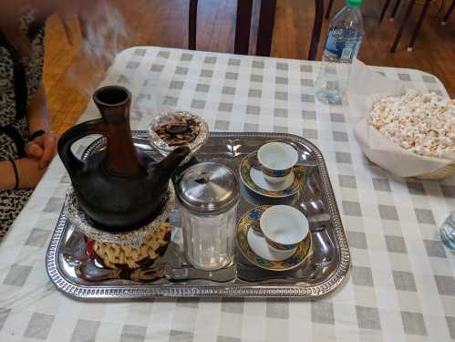 A silver tray with a steaming teapot, two cups, a sugar container, and a bowl of popcorn on a checkered tablecloth.