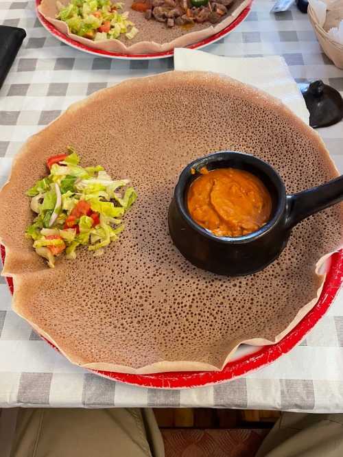 A large injera flatbread on a plate, accompanied by a small bowl of sauce and a side salad.