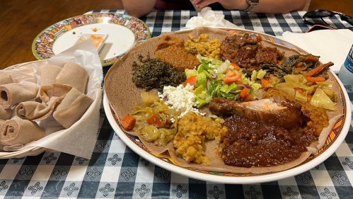 A large platter of Ethiopian food featuring injera, various stews, vegetables, and a side of rolled injera on a checkered tablecloth.