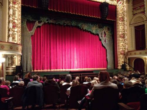 A theater stage with red curtains and decorative trees, filled with an audience of children and adults.