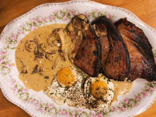 A plate featuring sliced beef, creamy mushroom sauce, and two fried eggs, garnished with herbs on a floral platter.