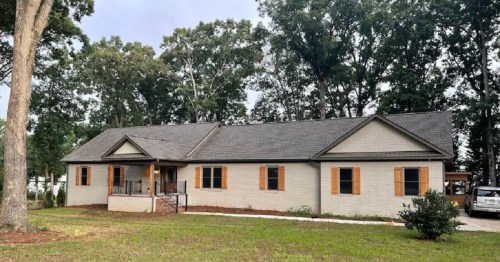 A single-story house with a gray roof, beige exterior, and wooden shutters, surrounded by trees and a grassy lawn.