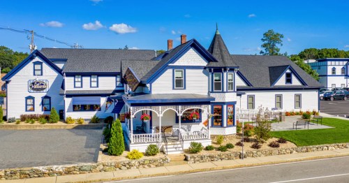 A charming blue and white Victorian-style building with a porch, surrounded by greenery and a clear blue sky.