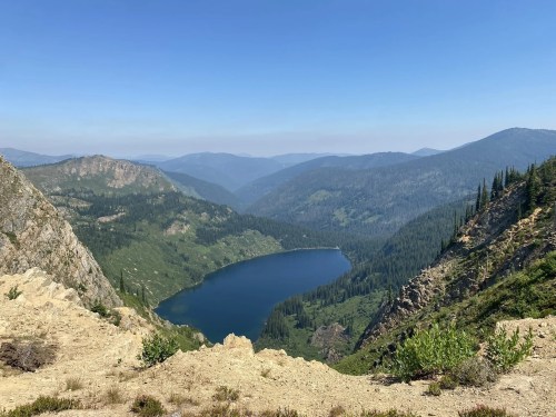 A serene mountain landscape featuring a blue lake surrounded by green forests and rolling hills under a clear sky.