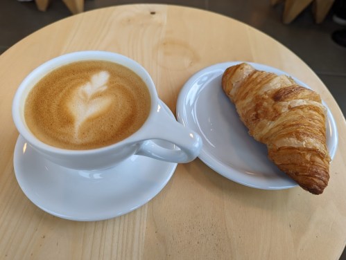 A cup of coffee with latte art next to a buttery croissant on a wooden table.