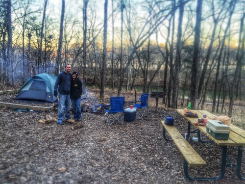 A couple stands together at a campsite with a tent, picnic table, and campfire setup in a wooded area.