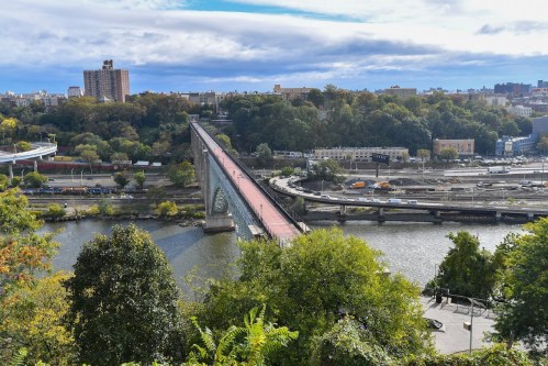 Aerial view of a bridge spanning a river, surrounded by trees and buildings under a partly cloudy sky.