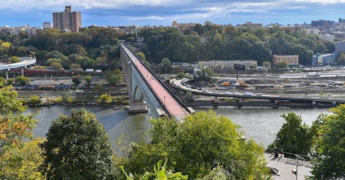 Aerial view of a bridge spanning a river, surrounded by greenery and urban buildings under a partly cloudy sky.