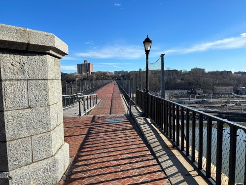 A scenic view of a brick walkway on a bridge, with a railing and buildings in the background under a clear blue sky.
