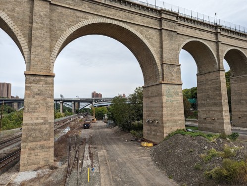 A large stone viaduct arching over a railway and highway, surrounded by greenery and urban structures in the background.