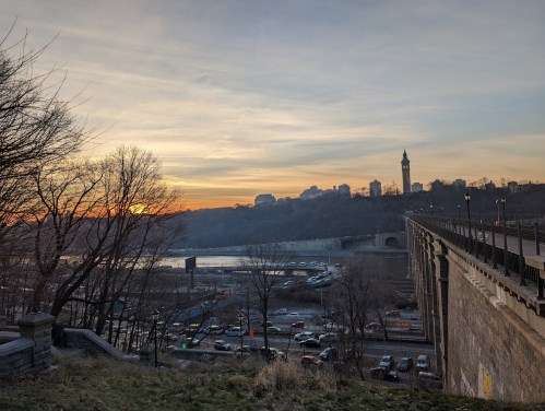 Sunset over a river with a bridge, trees in the foreground, and a city skyline in the background.