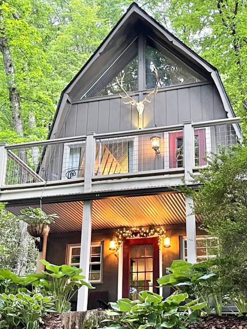 A charming A-frame cabin nestled in a forest, featuring a balcony, large windows, and decorative lights.