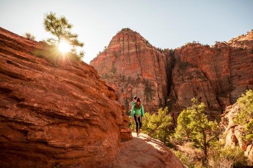 A hiker walks along a rocky path in a canyon, with sun shining behind towering red rock formations.