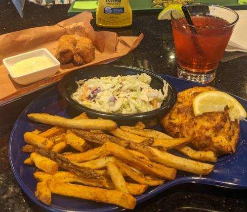 A plate with fries, coleslaw, a fish cake, and a dipping sauce, alongside a drink with a straw.