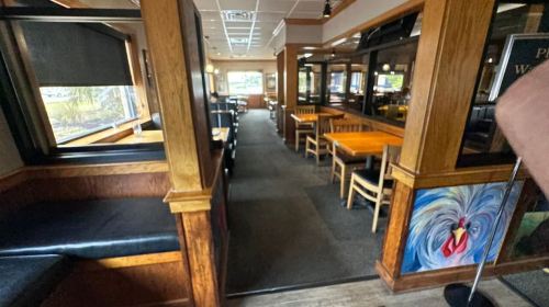 Interior of a restaurant featuring wooden tables, chairs, and a colorful mural on the wall. Natural light from windows.