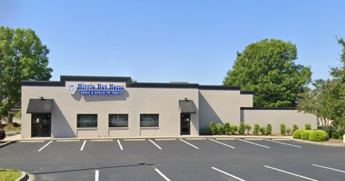 Exterior view of a single-story building with a sign reading "Hippie Hen House," surrounded by trees and a parking lot.