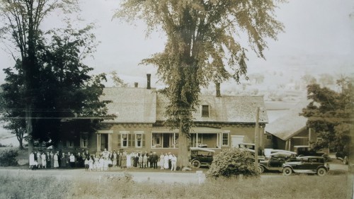 A historic black-and-white photo of a large group of people in front of a house, with vintage cars parked nearby.