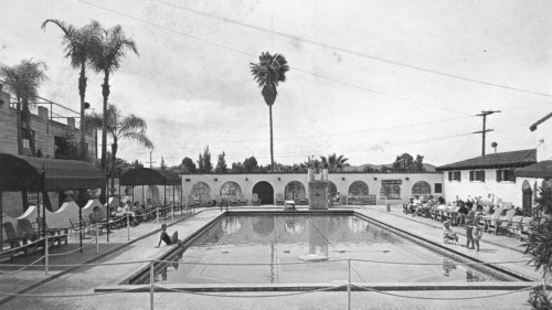Black and white photo of a pool area with palm trees, lounge chairs, and a decorative fountain.