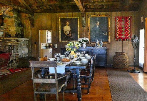 A rustic dining room with a long table set for a meal, featuring vintage portraits and a stone fireplace.
