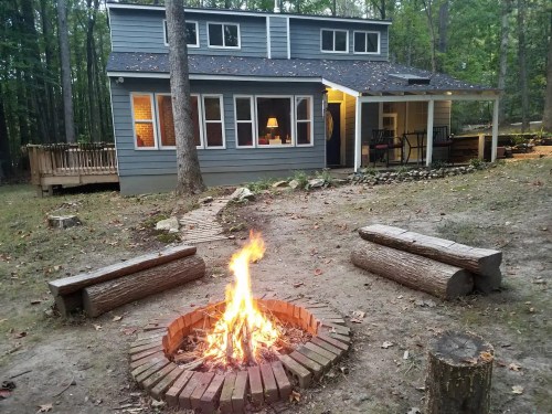 A cozy cabin in the woods with a fire pit and logs for seating, surrounded by trees and a peaceful outdoor setting.