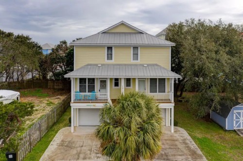 A two-story yellow house with a metal roof, surrounded by greenery and a palm tree in the front yard.