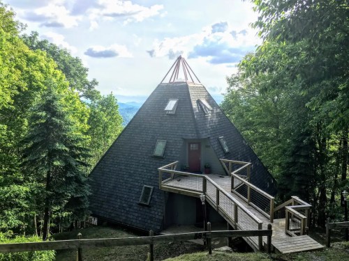 A unique pyramid-shaped house surrounded by trees, featuring a wooden deck and a scenic mountain view.