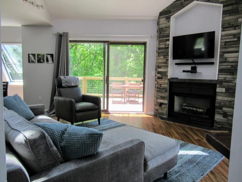 Cozy living room with a gray sofa, stone fireplace, and large windows overlooking a deck and greenery outside.