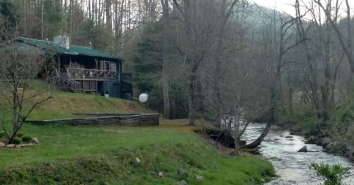 A cozy cabin by a stream, surrounded by trees and mountains, with a grassy area in front.