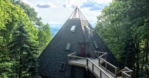 A unique triangular house with a wooden deck, surrounded by lush green trees and mountains in the background.