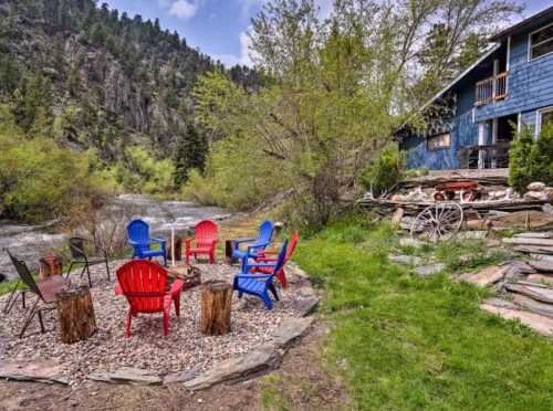 A cozy outdoor seating area with red and blue chairs around a fire pit, surrounded by greenery and a river.